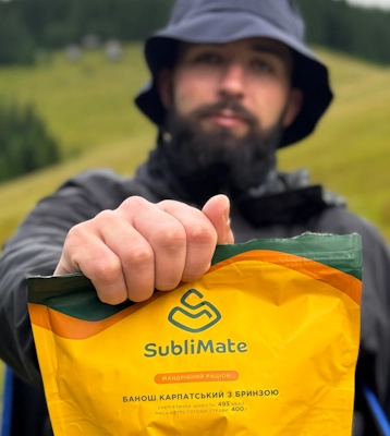 Турист у капелюсі тримає упаковку «Банош карпатський з бринзою» SubliMate на фоні зеленої гори