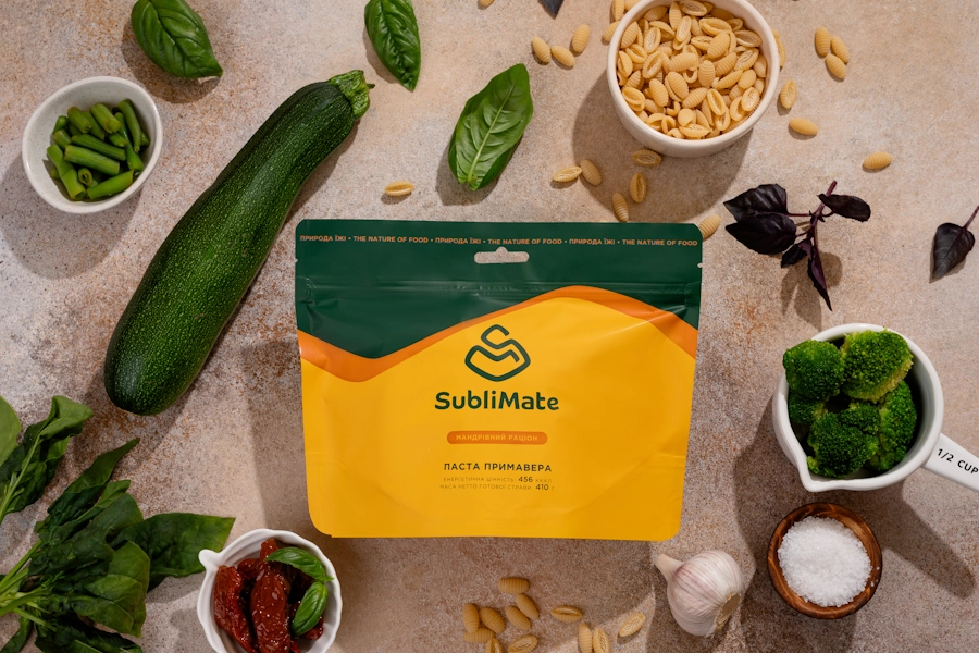 Яскраве жовто зелене паковання SubliMate оточене свіжими овочами і зеленню що входять до складу страви Примавера — композиція на кухонному фоні