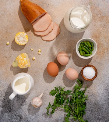 Складники страви SubliMate: яйця, молоко, копчена курка, зелень, кукурудза, квасоля, вершки та сіль на світлій поверхні.